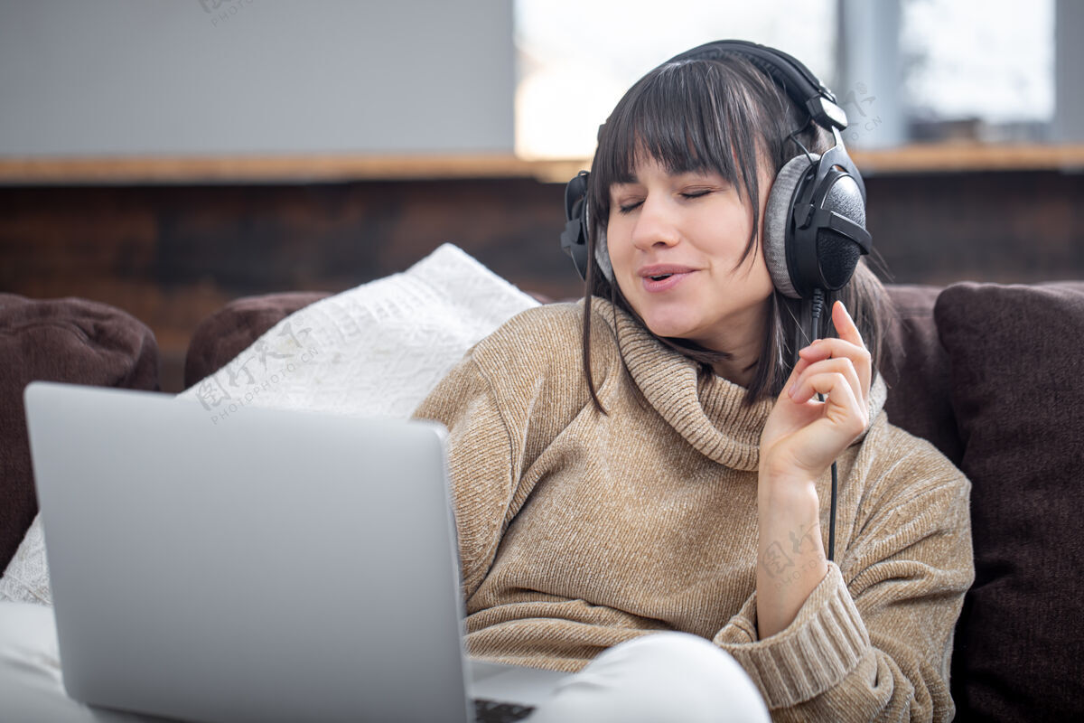 音乐美丽的女人戴着耳机在家里用笔记本电脑坐在沙发上听音乐听唱歌年轻