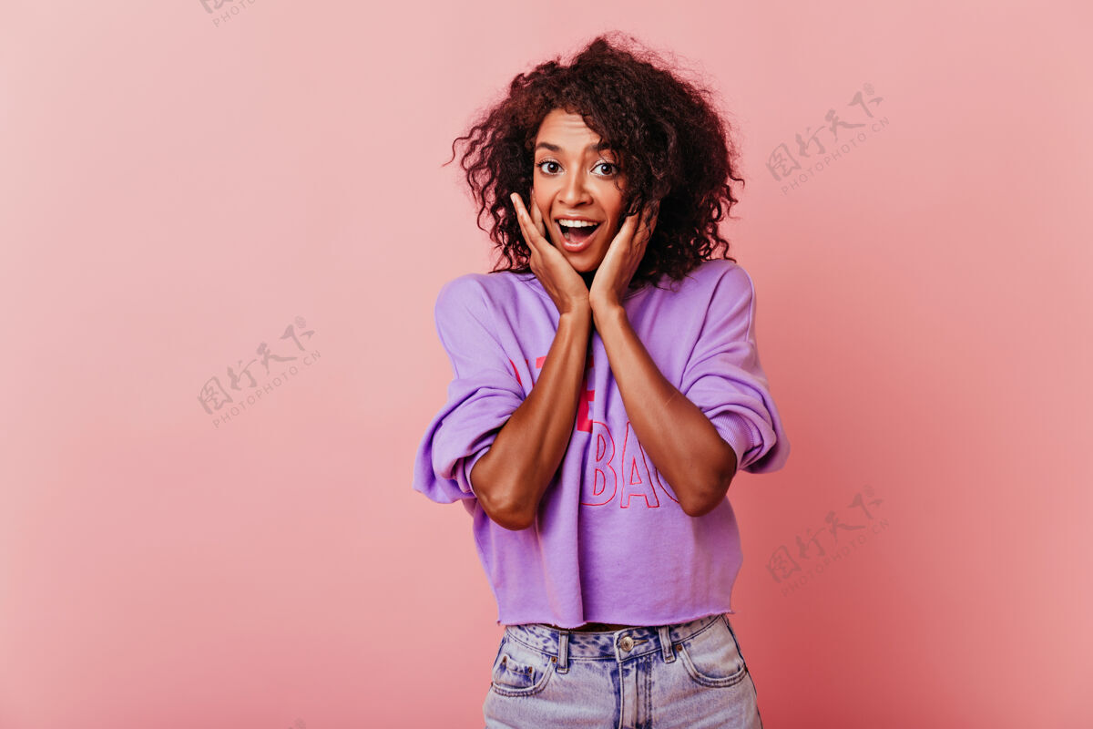 情感乐观的黑发女人抚摸着她的脸在粉彩上孤立的时尚非洲女孩的室内肖像粉色女性大笑