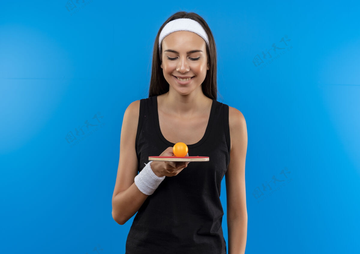 女孩微笑的年轻漂亮的运动女孩戴着头带和腕带拿着乒乓球拍看着球在蓝色的空间抱着腕带头带