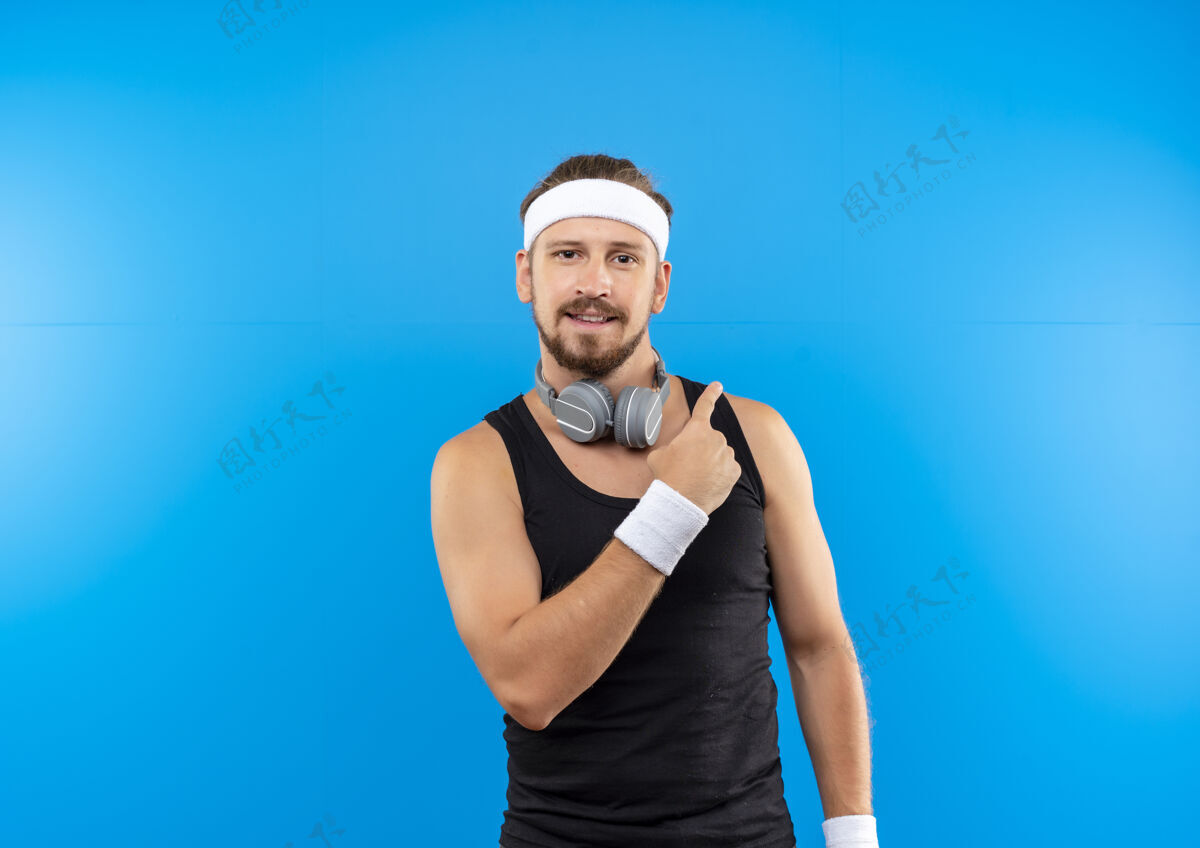 指向高兴的年轻帅气的运动型男子戴着头带和腕带 戴着耳机在脖子上指着隔离在蓝色空间的一边运动太空佩戴