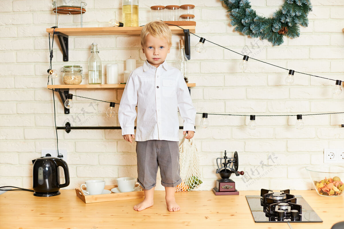 饭在时尚的斯堪的纳维亚厨房里 一头金发的可爱男婴光着脚站在木桌上 戴着圣诞花环 行为不端 却没有人看见他早餐肖像可爱