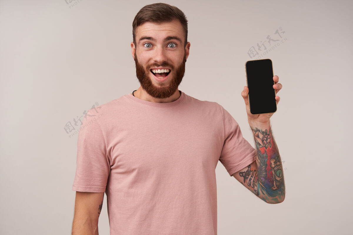 表情快乐的蓝眼睛美丽的胡须 纹身的男性穿着米色t恤 在白色的脸上摆姿势 开心地微笑着 举起手 里面有智能手机无胡子衣服男士