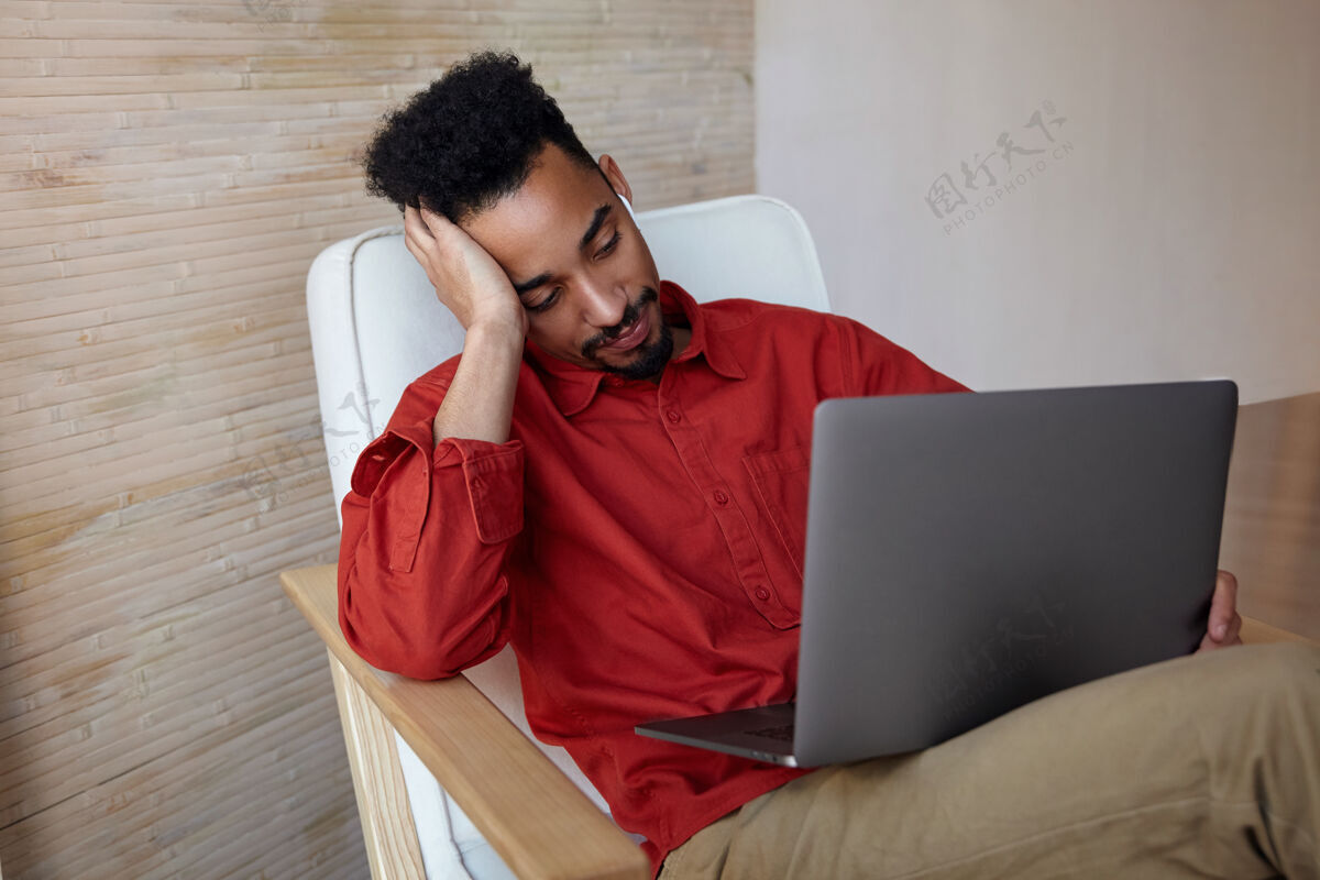 商务人士一幅室内肖像画 年轻的短发黑色男性坐在室内的椅子上 膝盖上放着笔记本电脑 头靠在抬起的手上 无聊地看着屏幕帅气椅子情绪