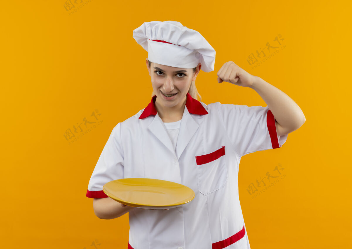 年轻年轻漂亮的厨师穿着厨师制服 戴着牙套 举着空盘子 举起拳头 孤立在橙色的空间里制服快乐厨师