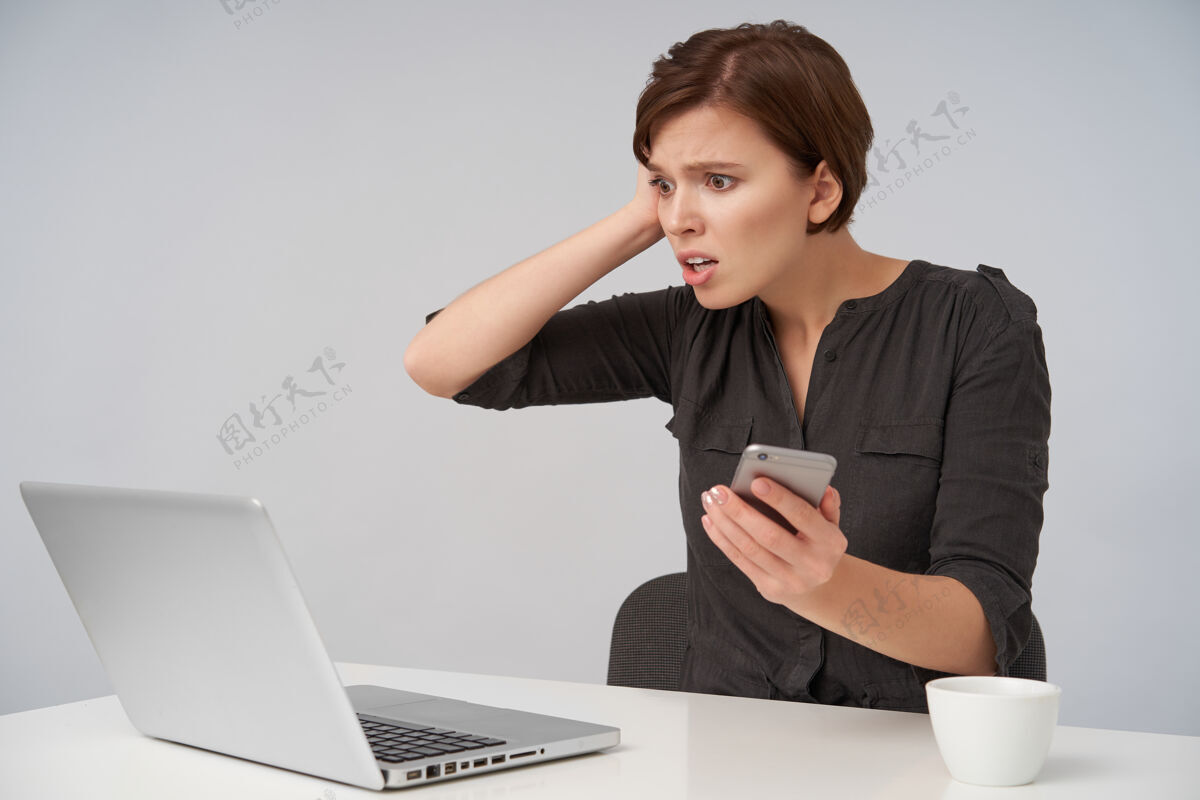 自然紧张的年轻漂亮的棕色头发的女性 短发时髦 举着手掌放在头上 圆圆的眼睛 惊奇地看着笔记本电脑的屏幕员工黑发坐着
