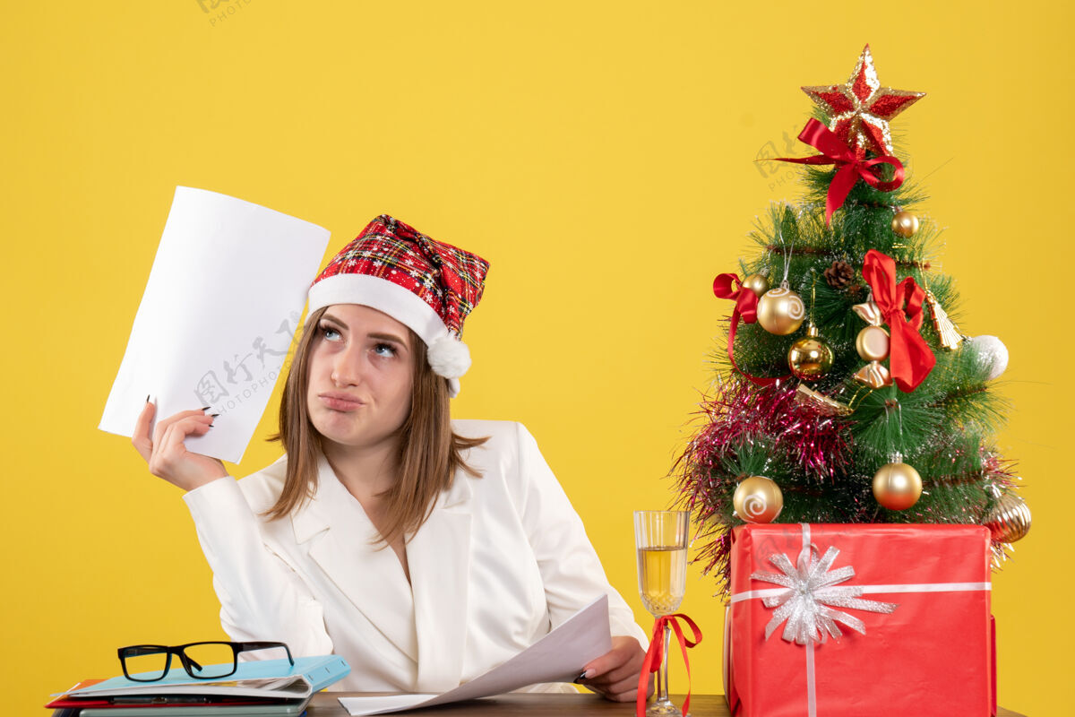 人前视图女医生坐在圣诞礼物拿着黄色背景上的文件微笑前面医生