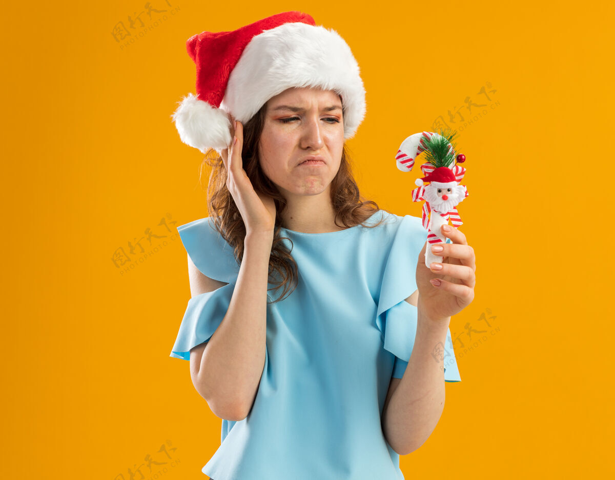帽子戴着蓝色上衣和圣诞帽的年轻女子拿着圣诞糖果手杖看着它困惑和不悦橙色看圣诞