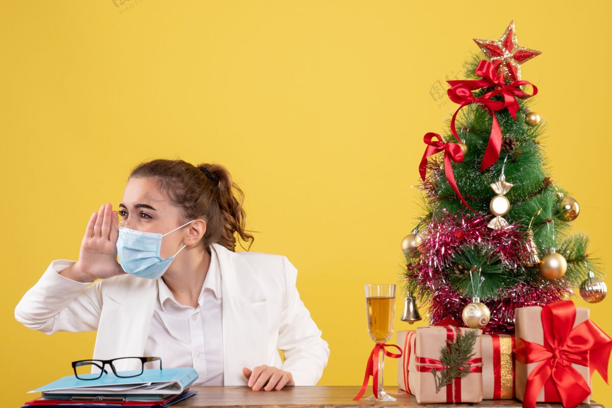 面具正面图女医生戴着无菌口罩坐在黄色背景上呼唤圣诞树和礼品盒人女性电话