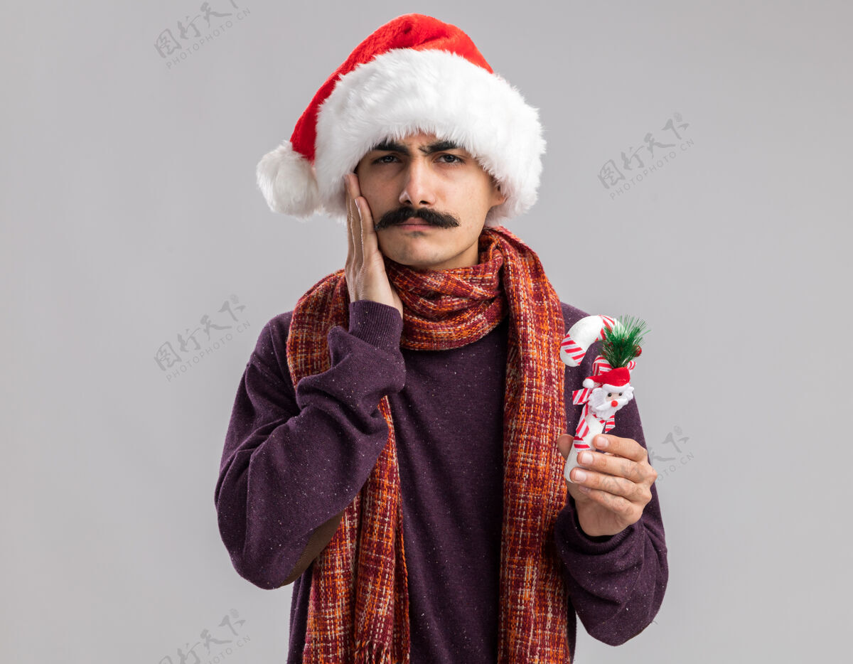 圣诞老人年轻的胡子男人戴着圣诞老人帽 脖子上围着暖和的围巾 手里拿着圣诞糖果手杖 看上去很困惑快乐年年轻