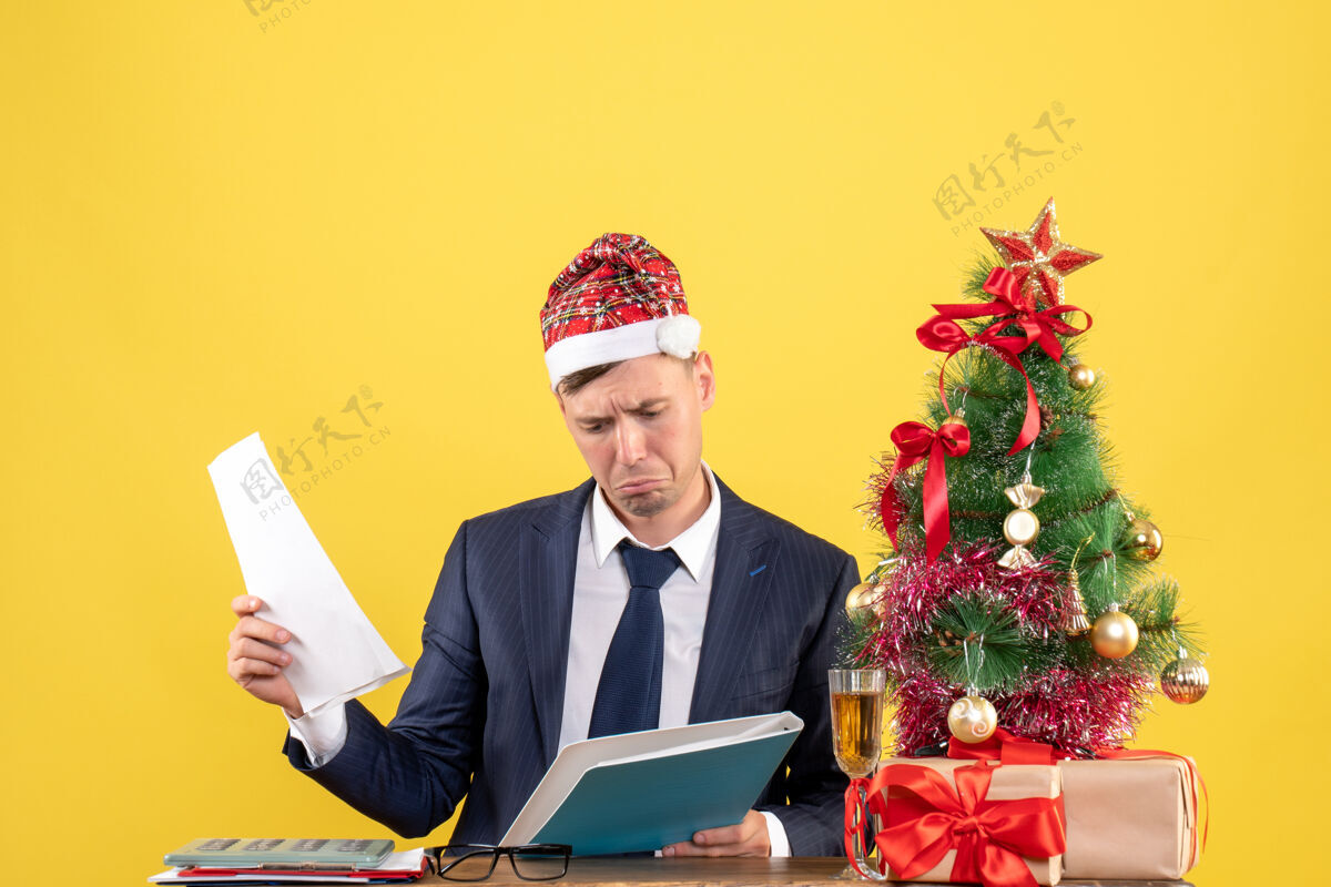 困惑困惑的商人坐在圣诞树旁的桌子前 呈现黄色公司黄色男性
