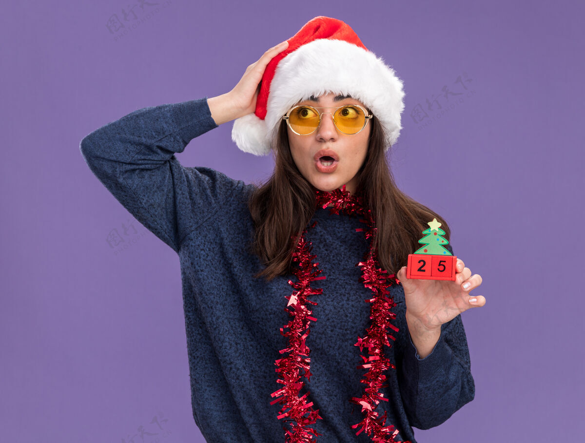 眼镜戴着太阳眼镜 脖子上戴着圣诞帽和花环的年轻白种人女孩手举圣诞树饰物 手放在头上 看着紫色背景上孤立的一面 还有复制空间快乐震惊花环