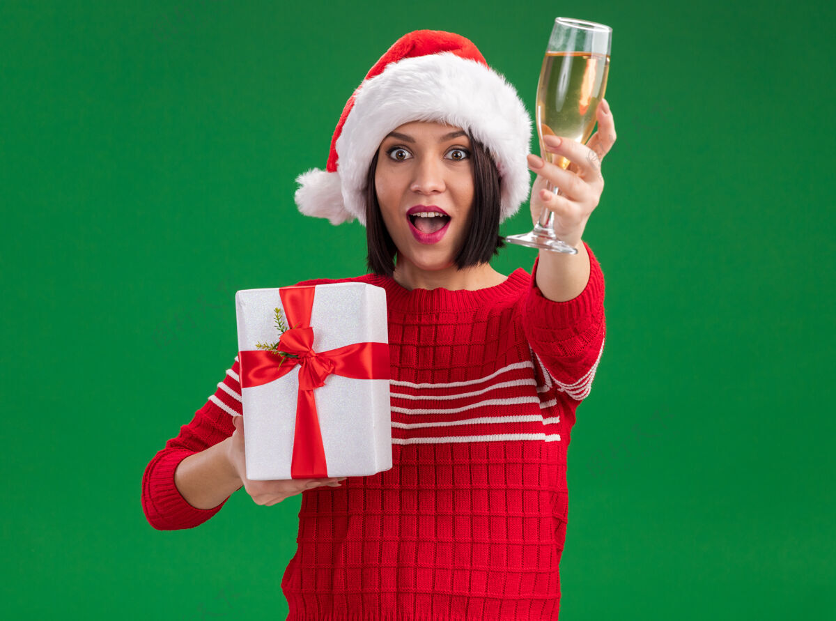 包装印象深刻的年轻女孩戴着圣诞帽拿着礼包 伸出一杯香槟隔离在绿色的墙上圣诞老人年轻穿着