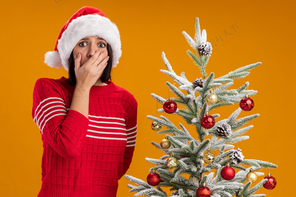 树一个戴着圣诞帽的年轻女孩站在装饰好的圣诞树旁 手放在嘴上 另一个站在后面 看着隔离在橙色背景上的摄像机穿看帽子