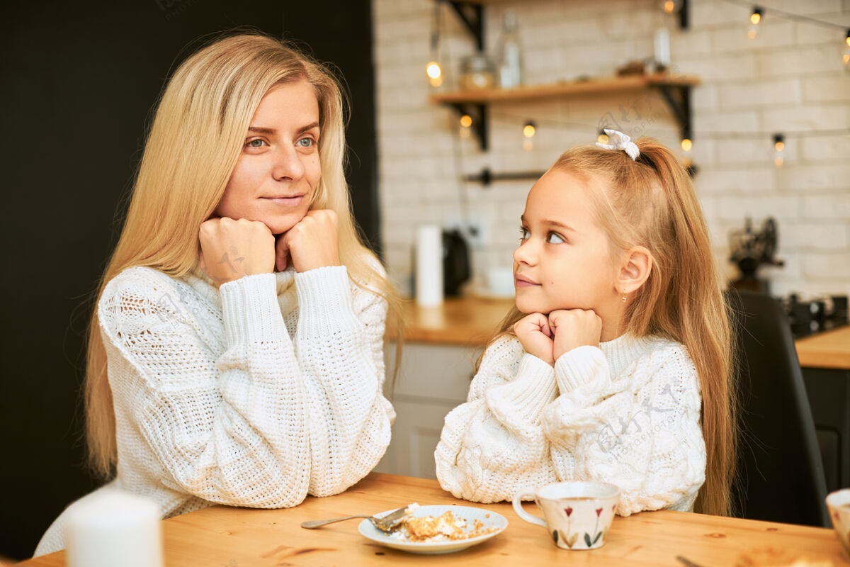 女儿漂亮的金发女青年和漂亮的女儿都穿着舒适的毛衣在厨房里吃早餐 坐在餐桌旁喝茶 吃蛋糕 手放在下巴下面妈妈蛋糕年轻