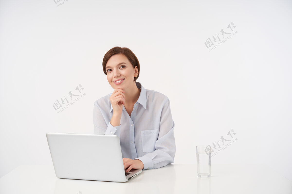 办公室积极的年轻棕色眼睛的黑发女性 随意的发型keepig抬起手放在下巴上 愉快地看着旁边 带着迷人的微笑 坐在白色的椅子上休闲好情绪