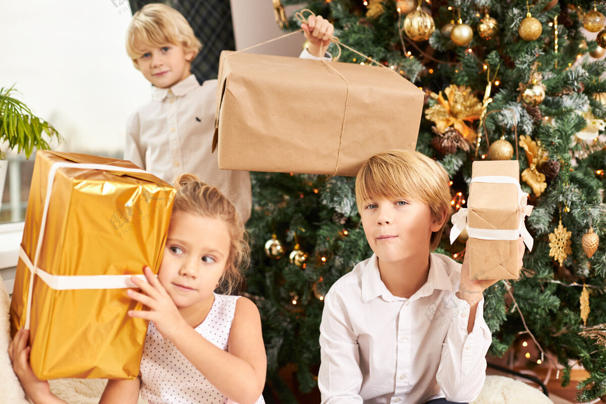 庆祝三个可爱的兄弟姐妹坐在装饰好的新年树旁 手里拿着圣诞礼物的盒子 感到不耐烦 神情好奇快乐的童年 快乐的节日圣诞节孩子幸福