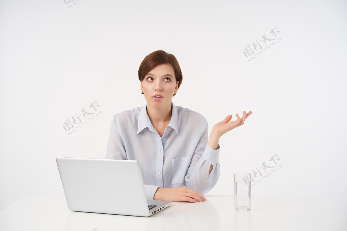 笔记本电脑年轻的棕色眼睛短发黑发女子坐在桌旁 手里拿着笔记本电脑 撅着嘴 抬起手掌 在白色的桌子上摆姿势情绪玻璃蓝色