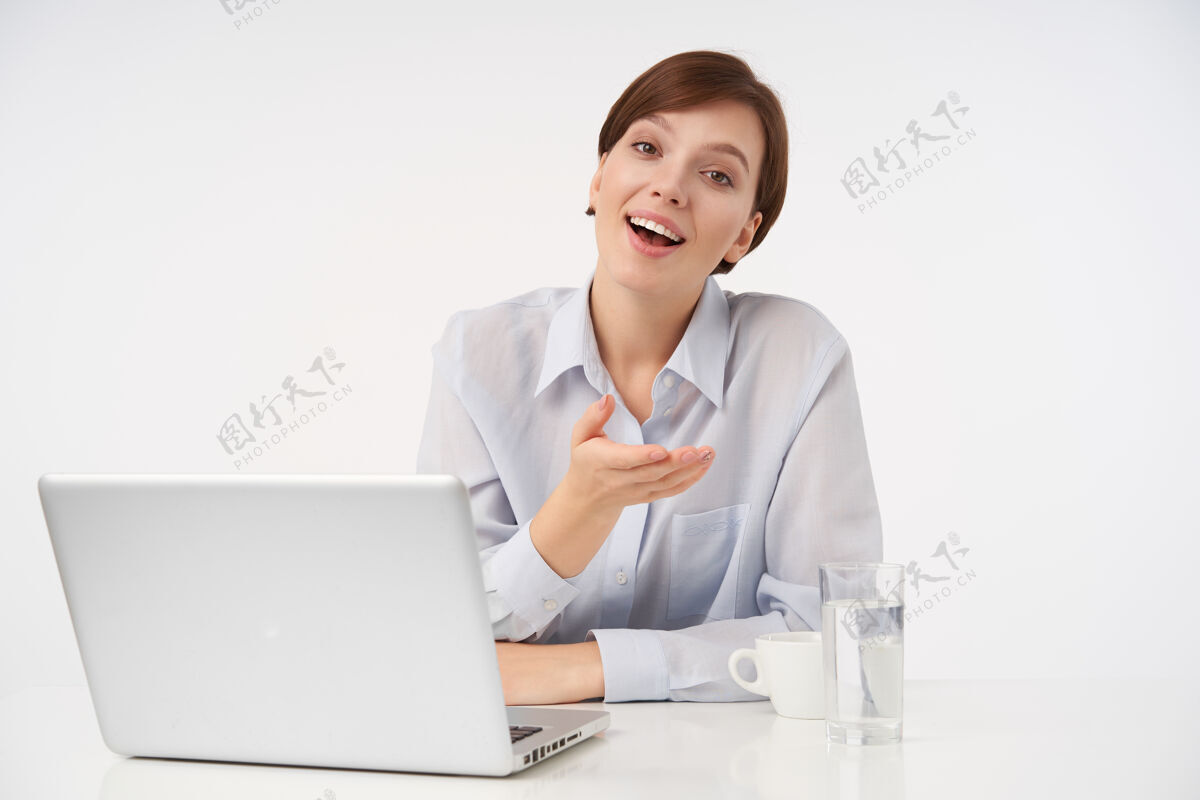 坐着快乐的年轻棕色眼睛短发女士的室内肖像 随意的发型 手持笔记本电脑在现代办公室工作 抬起手掌 愉快地微笑优雅办公室自然