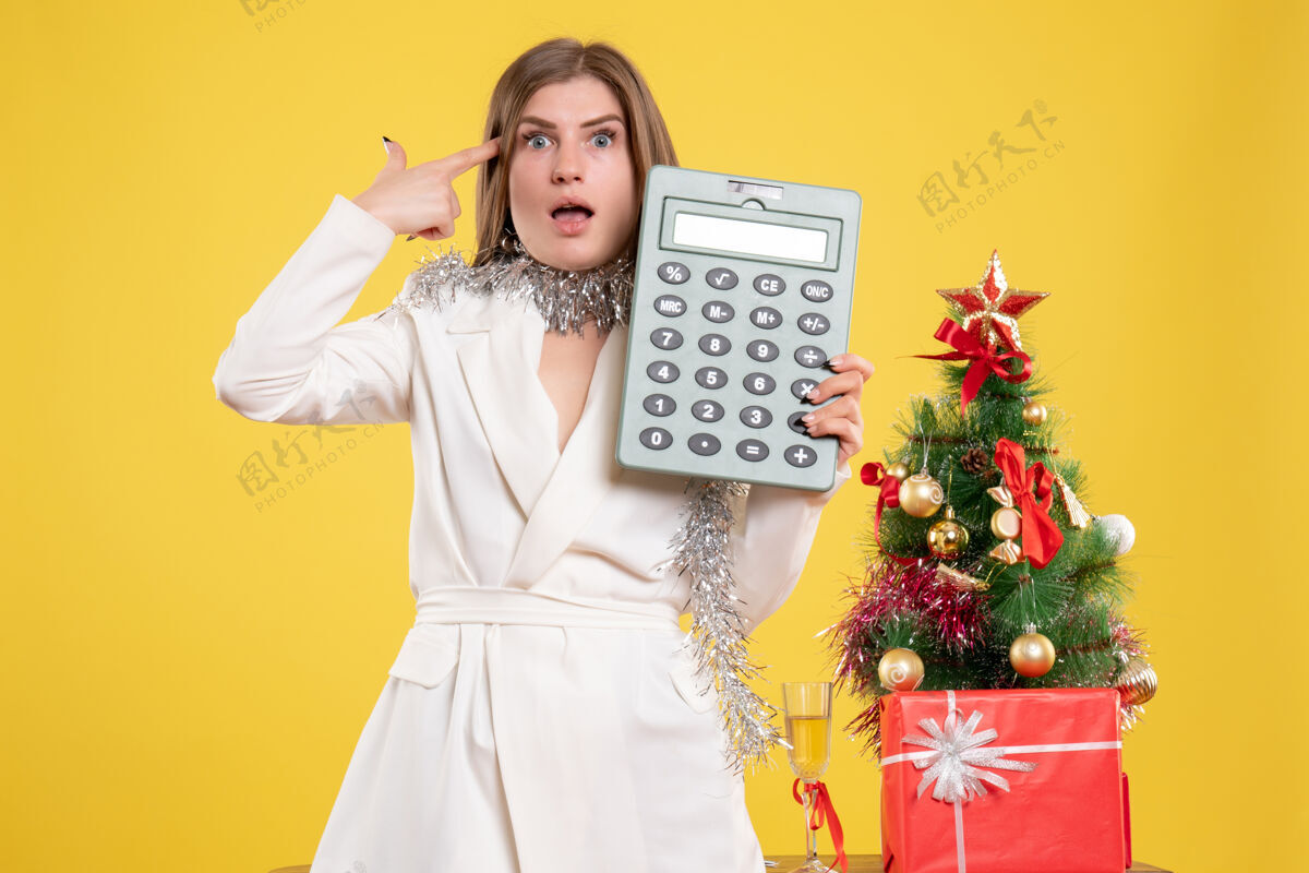 肖像正面图女医生站着拿着计算器 背景是黄色的圣诞树和礼品盒护士女医生女