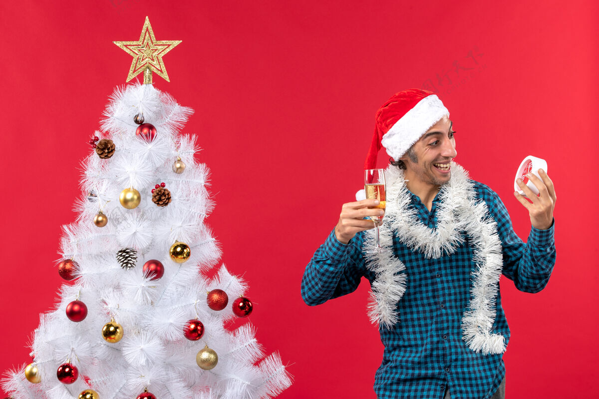 年轻人带着圣诞老人帽子 拿着一杯葡萄酒 微笑着 情绪激动的年轻人 在红地毯上的圣诞树旁的钟声前摇摇晃晃酒人年轻人