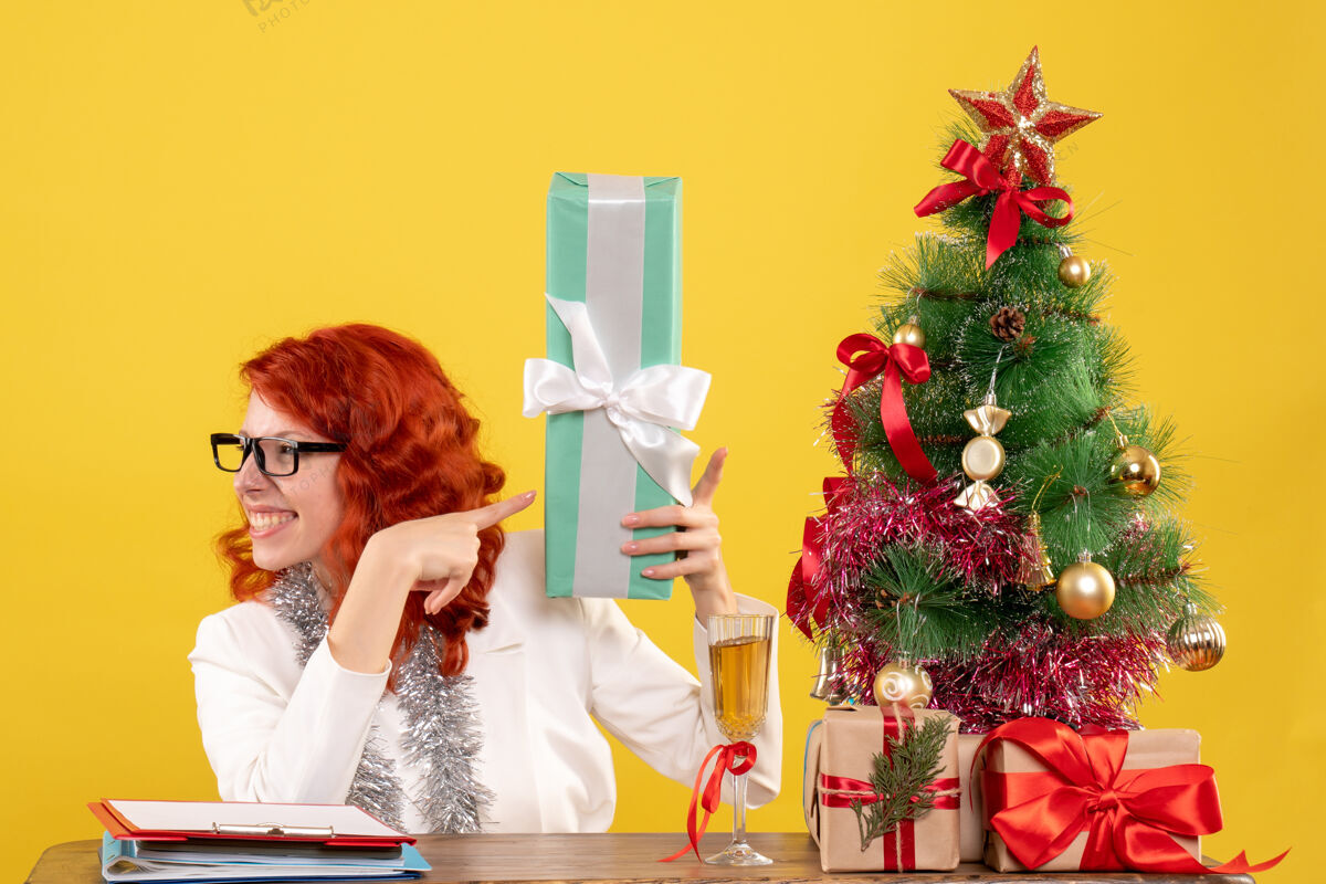 微笑正面图：女医生坐在黄色的桌子上 手里拿着圣诞礼物和圣诞树人新郎礼物