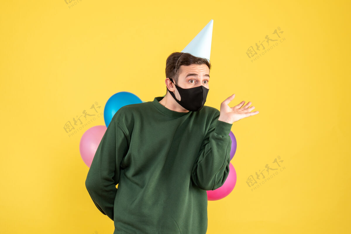 脸前视图迷惑了一个戴着派对帽 站在黄色地面上的彩色气球的年轻人五颜六色商人男子气概