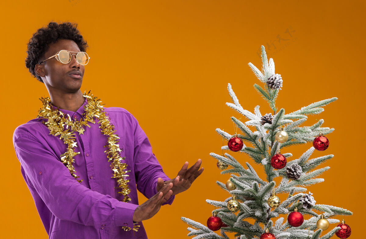 戴着一个戴着眼镜 脖子上戴着金箔花环的年轻非裔美国人站在装饰过的圣诞树旁 看着它在橙色的墙上做着一个孤立的手势金属片男人未发布