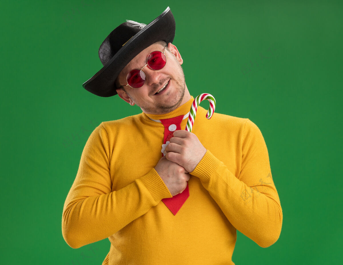 高领毛衣穿着黄色高领毛衣 戴着眼镜 打着滑稽的红领带 戴着黑色帽子 手里拿着糖果棒 脸上带着幸福的笑容站在绿色的墙上年轻人圣诞节手杖