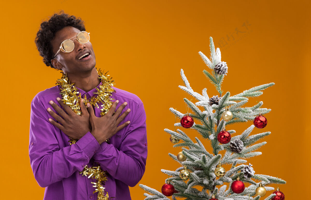 周围令人印象深刻的年轻非裔美国人戴着眼镜 脖子上戴着金箔花环 站在橙色背景的装饰圣诞树旁脖子装饰近