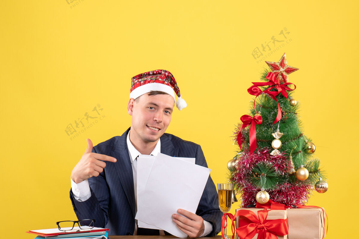 团队正面图：快乐的男人指着坐在圣诞树旁桌子上的文件和黄色的礼物礼物快乐文件