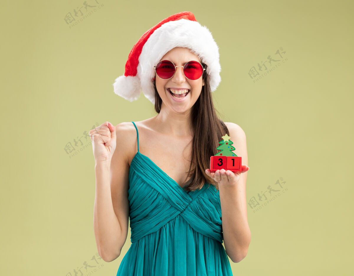 快乐戴着太阳眼镜 戴着圣诞帽的快乐白人女孩握紧拳头 手持圣诞树饰物圣诞快乐拳头橄榄