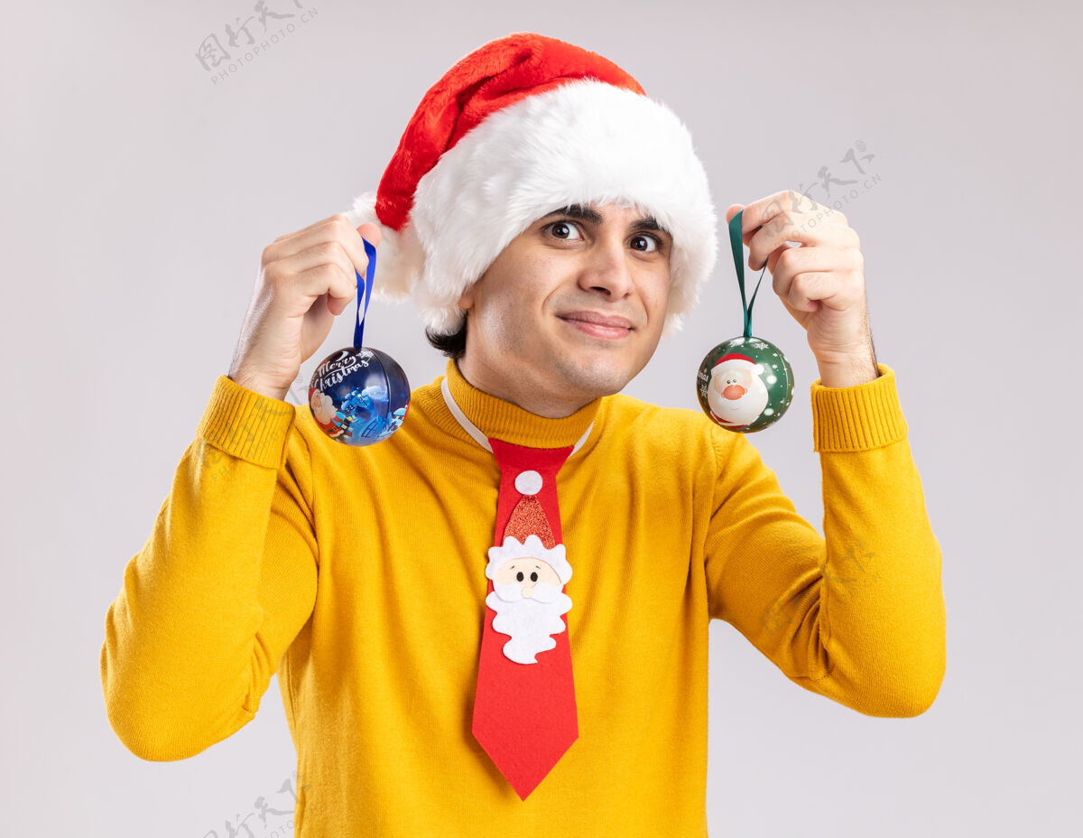 球穿着黄色高领毛衣 戴着圣诞老人帽的年轻人举着圣诞球站在白色的墙上 开心而积极地微笑着男人积极帽子
