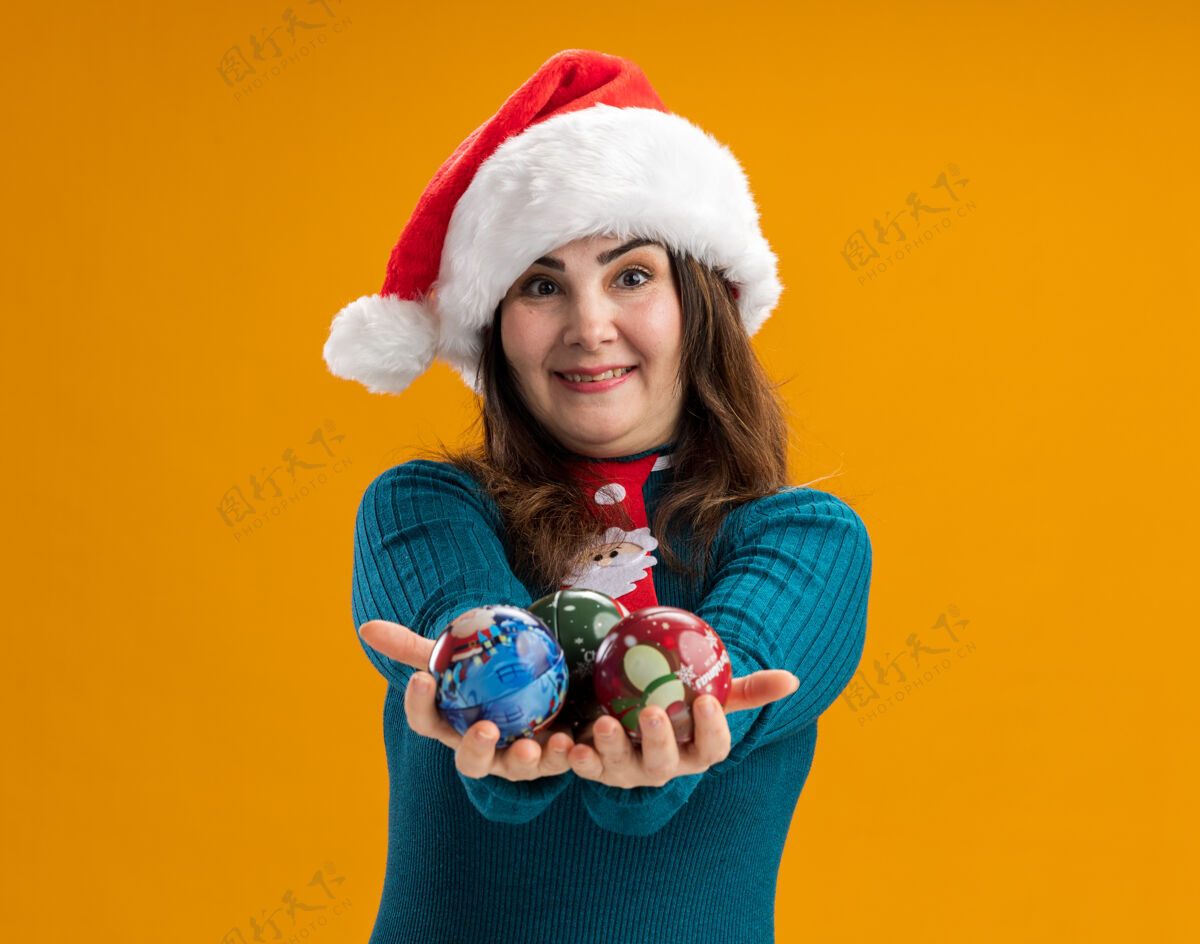 成人令人印象深刻的成年白人妇女与圣诞帽和圣诞领带举行的玻璃球饰品孤立的橙色背景与复制空间快乐背景帽子