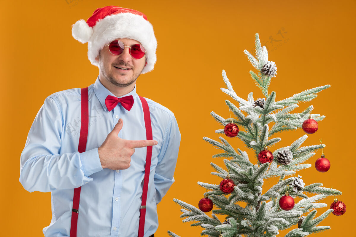 手指快乐的年轻人戴着吊带领结 戴着圣诞帽 戴着红眼镜 站在圣诞树旁 用食指指着圣诞树 在橙色的墙上微笑着圣诞节年轻人手指