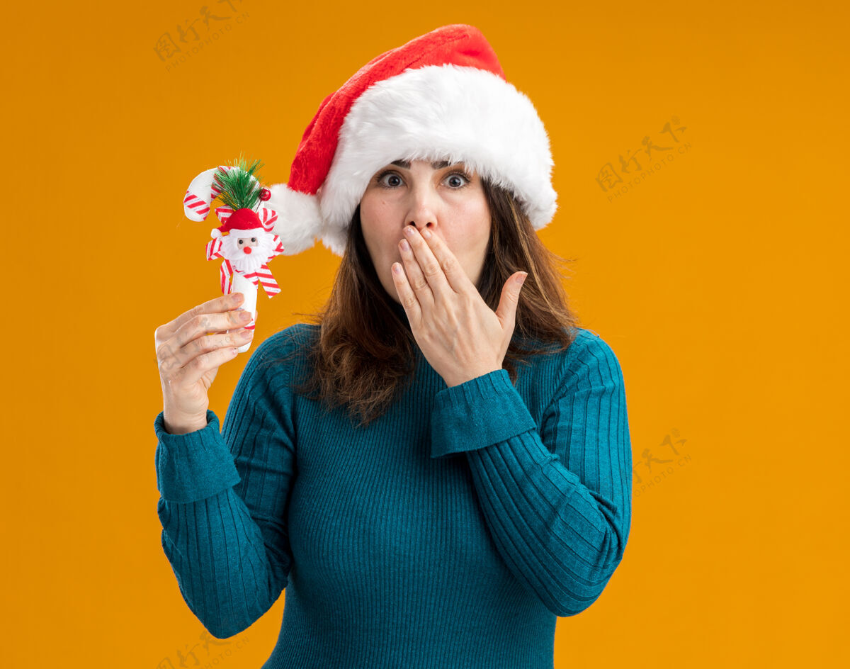 震惊震惊的成年白人妇女戴着圣诞帽 把手放在嘴上 拿着糖果棒孤立地放在橙色背景上 留有复制空间嘴快乐帽子