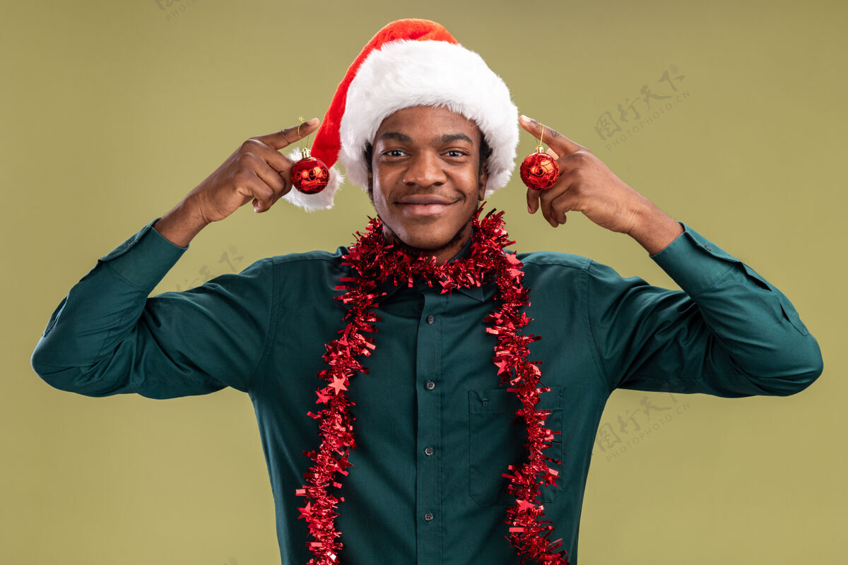 圣诞老人快乐的非洲裔美国人戴着圣诞帽 戴着花环 拿着圣诞球 微笑着站在绿色的墙上男人花环圣诞节