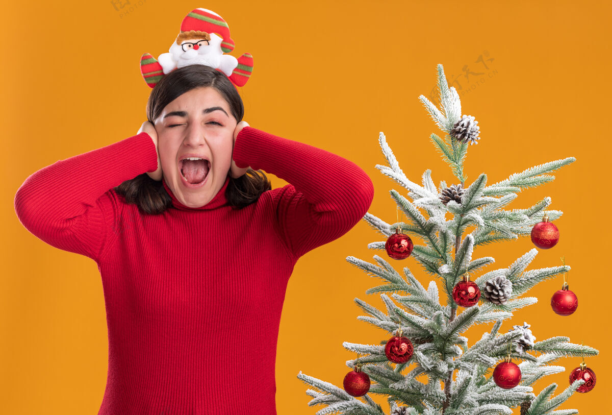 头带穿着圣诞毛衣的年轻女孩戴着滑稽的头带疯狂地高兴地闭上耳朵双手站在橙色墙上的圣诞树旁站着毛衣穿着