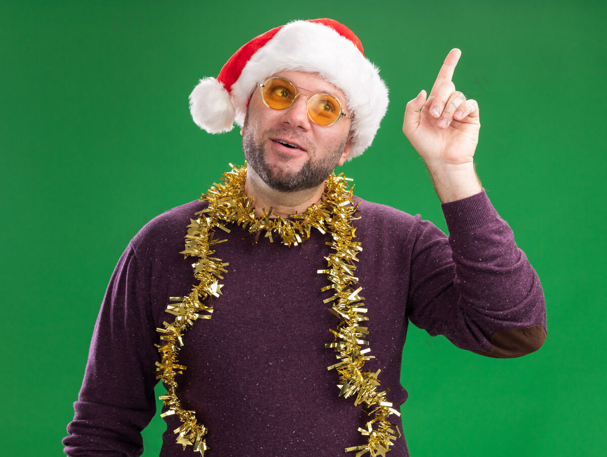 圣诞老人令人印象深刻的中年男子戴着圣诞帽 脖子上戴着金箔花环 戴着眼镜 一边看着一边指着绿色的背景金属丝眼镜周围