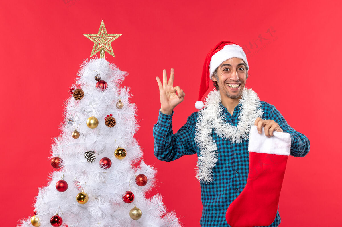 袜子兴奋的年轻人戴着圣诞老人的帽子 穿着蓝色的衬衫 手里拿着圣诞袜子圣诞树男人年轻人