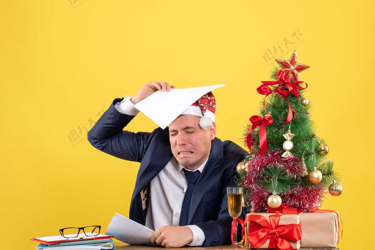 摩托板失望的男人坐在圣诞树旁的桌子前 黄色的礼物失望的男人商人礼物