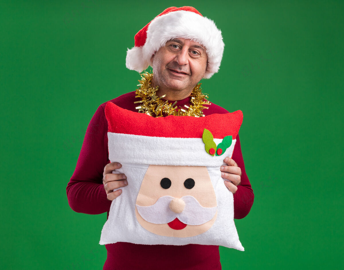站着快乐的中年男人戴着圣诞老人帽 脖子上戴着金属丝 抱着圣诞枕头 面带微笑地看着相机 站在绿色的背景上背景微笑金属片