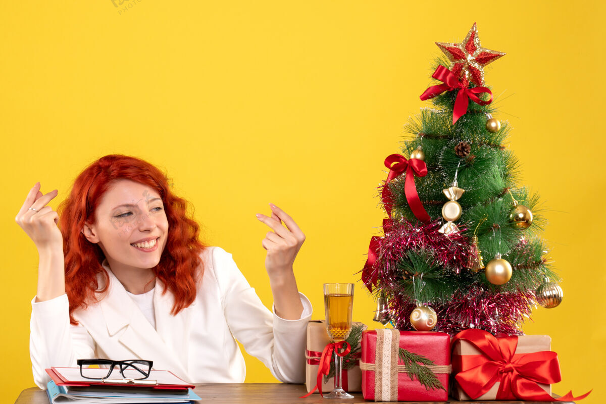 插花正面图：女医生坐在桌子后面 黄色背景上放着圣诞礼物和圣诞树花快乐礼物