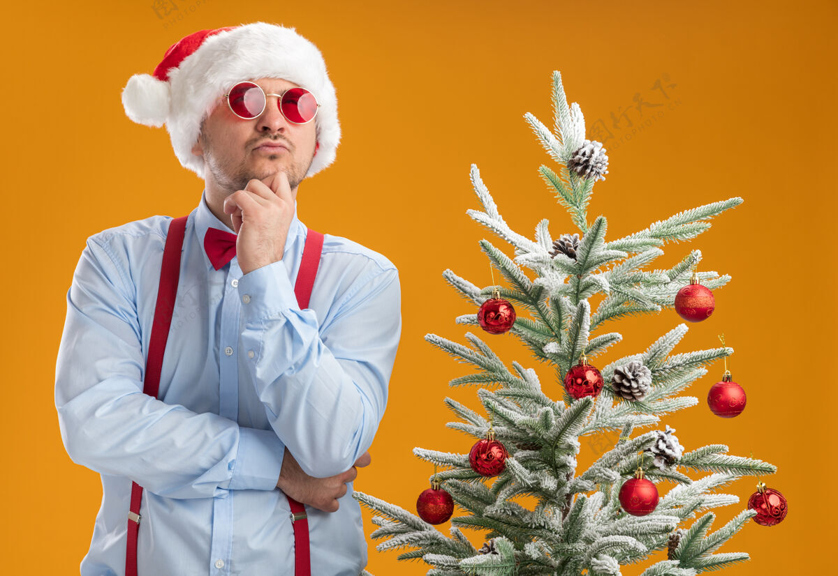 困惑一个戴着圣诞帽 戴着红眼镜 系着吊带领结的年轻人站在橙色墙上的圣诞树旁 困惑地抬头望着蝴蝶结穿着圣诞节