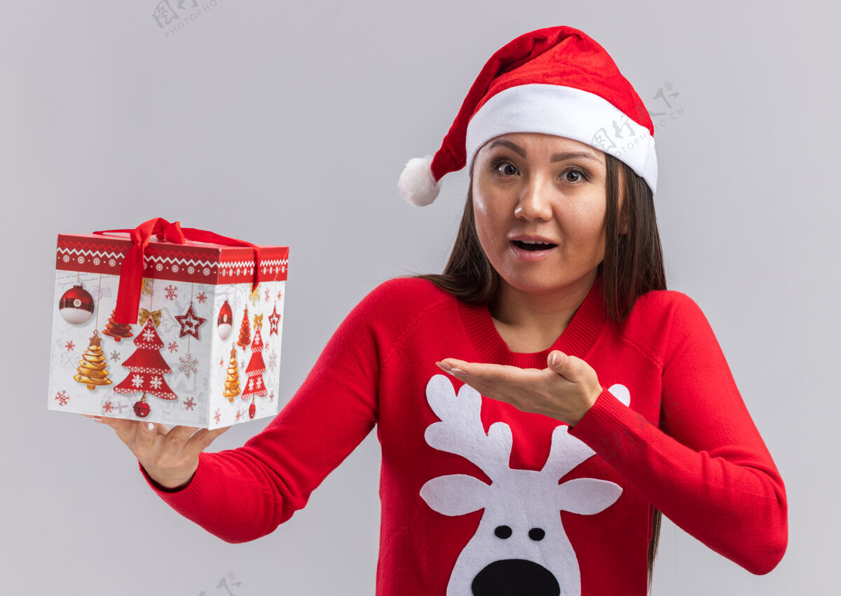 惊喜惊讶的年轻亚洲女孩戴着圣诞帽拿着毛衣 指着隔离在白墙上的礼盒年轻点女人