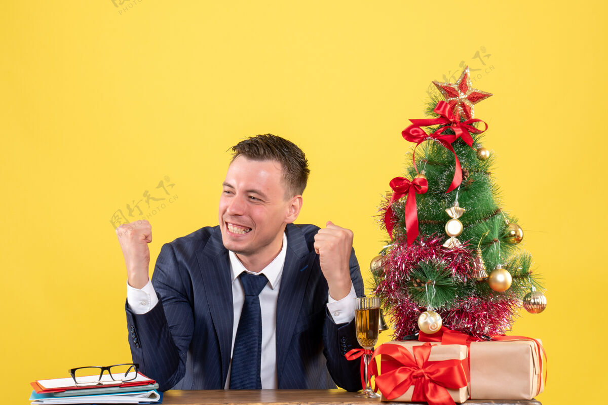 男人前视图微笑的人显示获胜的姿态坐在桌旁圣诞树和礼物黄色桌子公司男性