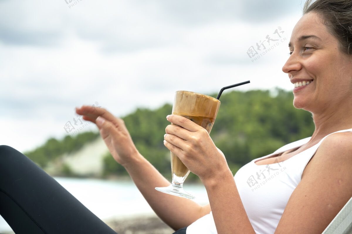 商店微笑的高加索妇女拿着咖啡饮料在沙滩上与泡沫和喝稻草与山露台水海洋