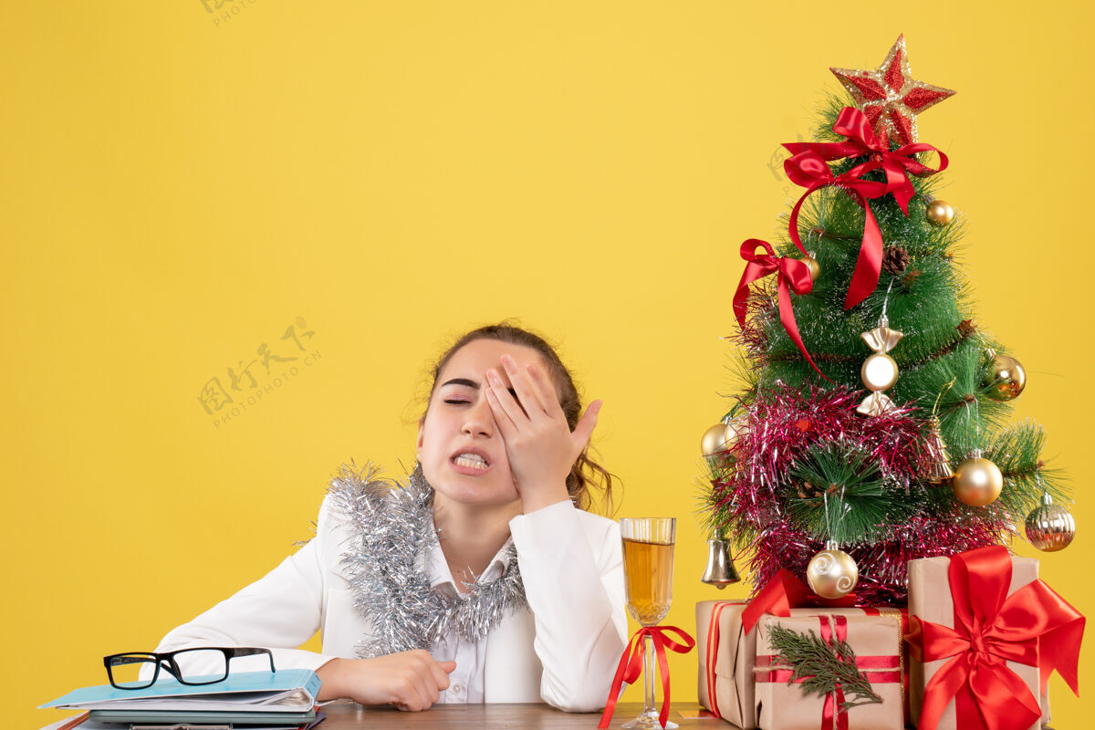 圣诞节正面图：女医生坐在桌子后面 黄色背景上有圣诞树和礼品盒前面快乐情绪