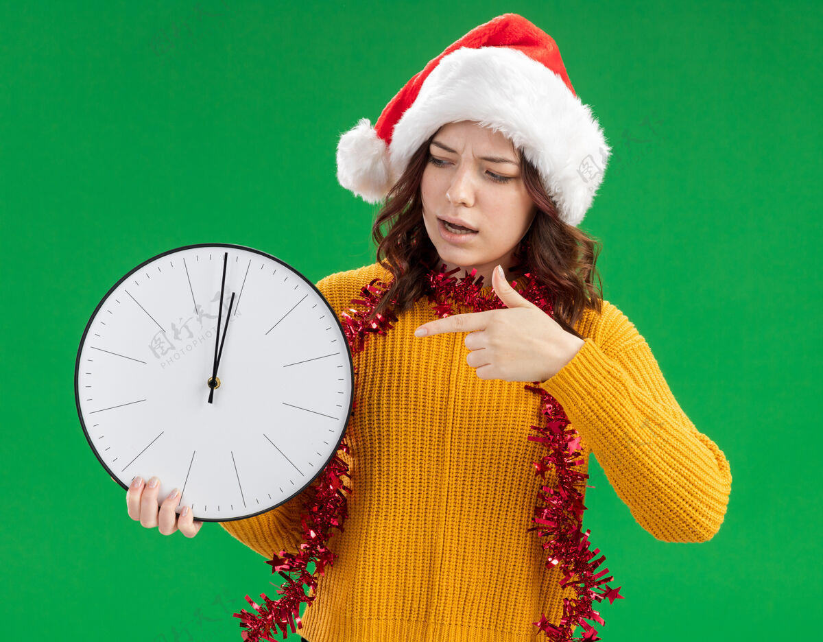 快乐无知的年轻斯拉夫女孩 戴着圣诞帽 脖子上戴着花环 拿着并指着绿色背景上的时钟 还有复制空间围着圣诞老人新