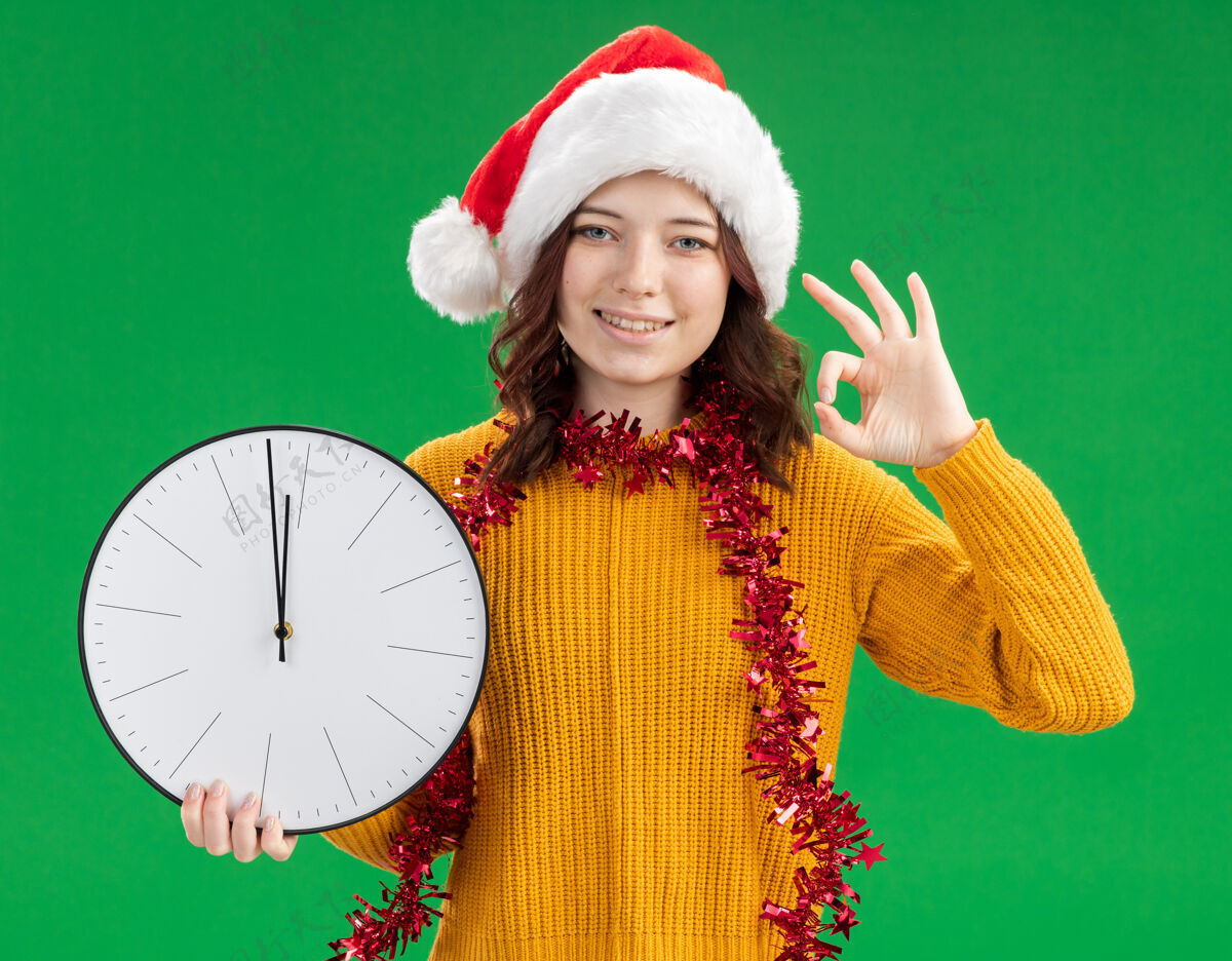 圣诞节微笑着的年轻斯拉夫女孩 戴着圣诞帽 脖子上戴着花环 拿着时钟 打着ok的手势 绿色背景上有空白女孩微笑快乐