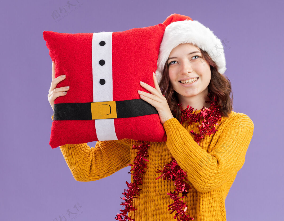 抱年轻的斯拉夫女孩戴着圣诞帽 脖子上戴着花环 抱着装饰好的枕头 在紫色的背景下 看着孤立的复制空间脖子圣诞老人女孩